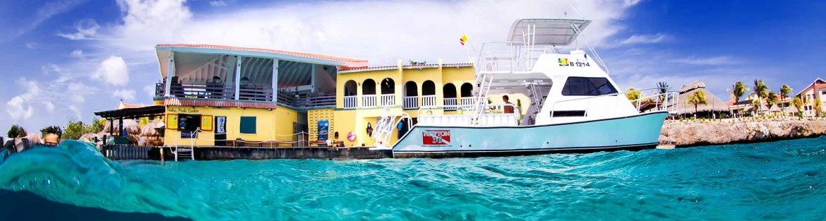 Blue Caribbean water in the foreground with a blue sky. In the middle of the photo is a bright colored building, with a boat docked out front, held by Sapias Holding B.V.