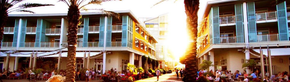 Ministry of District Administration, Tourism and Transport. Two multistory buildings on the street with a lot of people on the sidewalks of buildings. Sun peaking past a palm tree in the center of the photo, donw the alley.