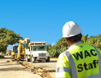 A worker with a hard hat and WAC Staff reflector vest looks on at an excavator and dump truck at work.