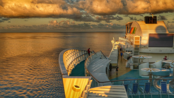 Upper deck of a cruise ship with people walking along it. Puffy clouds and water to the left and behind.