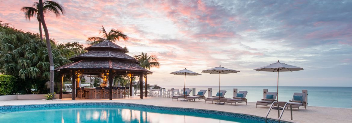 Blue Waters Resort and Spa - Antigua. A view of one of their pools with a tiki bar and poolside lounges and umbrellas; ocean and sky behind.