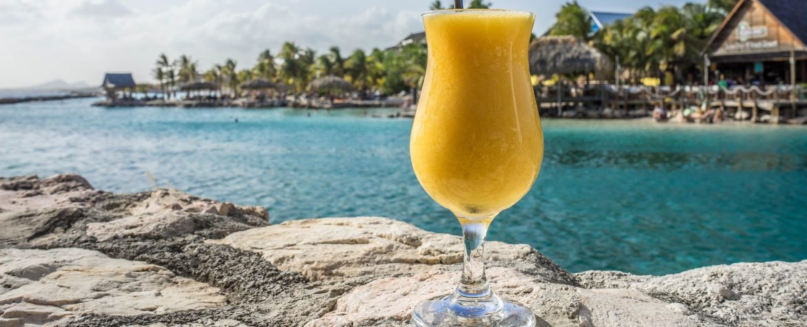 Caribbean Tourism Organization. A drink sitting on a rock with water, palm trees and tiki buildings behind. A tropical setting.