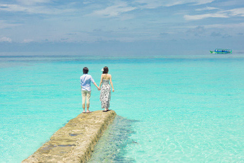 Caribbean Tourism Organization. A beach scene with two people holding hands and standing on a cement dock with algae from being submerged. Bright blue water all around with a boat and clouds in the distance.