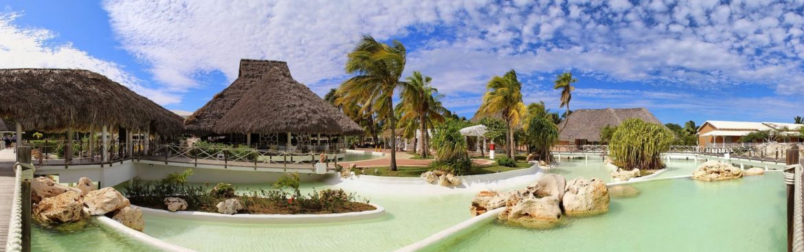 Ministry of Tourism & Investment, Antigua and Barbuda. Palm trees, pool, tiki hut and blue sky with white clouds.