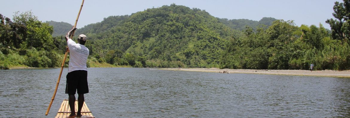 A man steering a raft on water with a pole and mountains in the background.