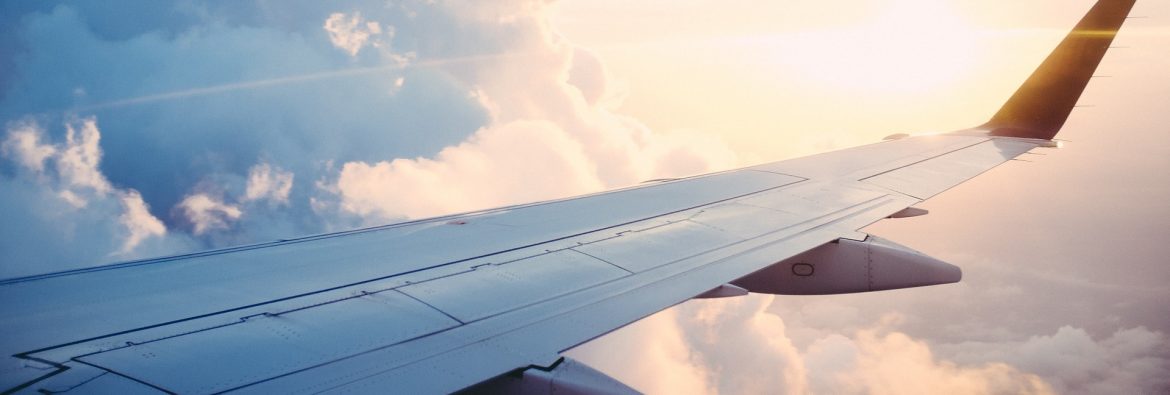 An airplane wing in the sky with clouds and sun shining behind.