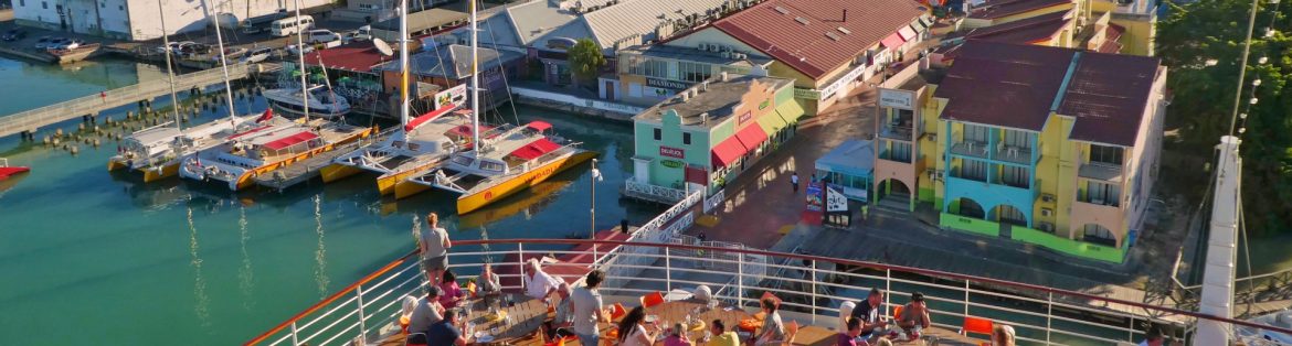 View of a Caribbean town from the back of a ship with people dining on the deck and the town beyond.
